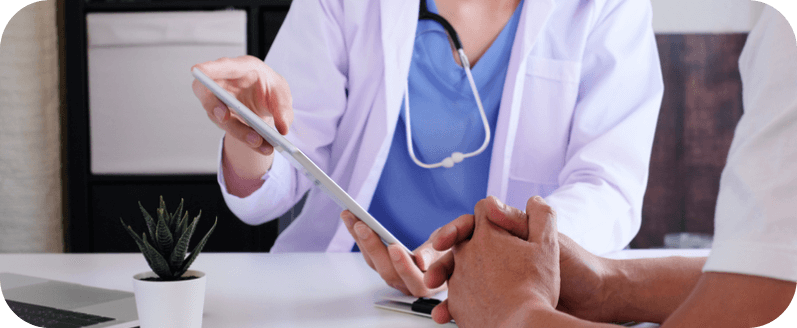 A doctor wearing a white coat holds a tablet in hand while discussing medical information with a patient, providing guidance and explanation.