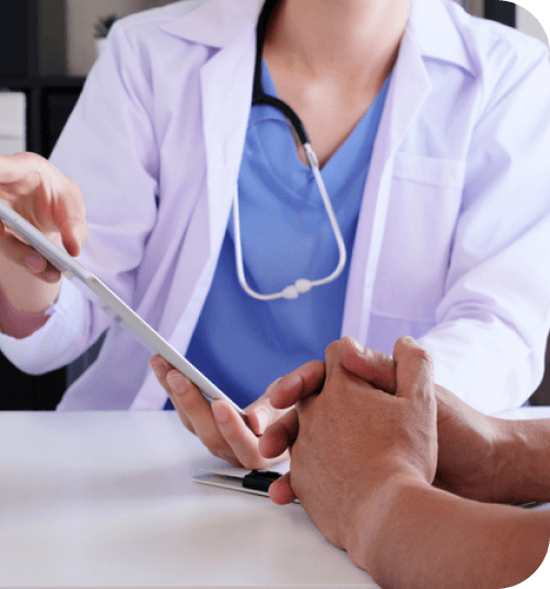 A doctor wearing a white coat and stethoscope holds a tablet while discussing with a patient.