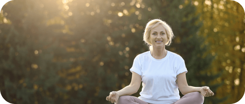 A smiling senior woman in a white t-shirt practicing meditation outdoors, sitting cross-legged on a green lawn with trees in the background, basking in soft sunlight.