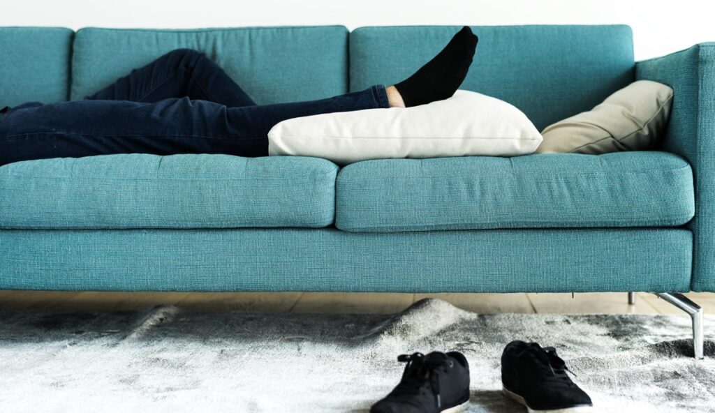 A person with their legs propped up on a pillow, resting on a teal couch. Their feet are in black socks, and a pair of shoes is on the floor beside the couch.