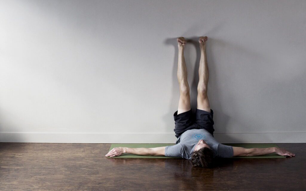 A male yoga instructor does the Viparita Karani or the Legs Up the Wall pose in an indoor yoga studio.