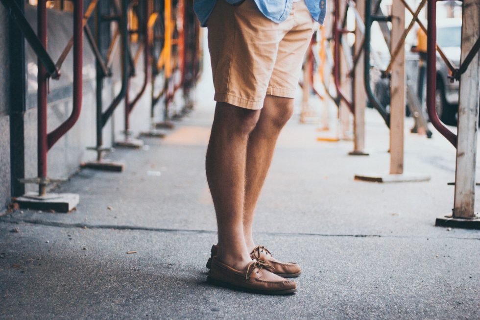 A man wearing khaki shorts and brown boat shoes stands on a sidewalk with construction scaffolding in the background.