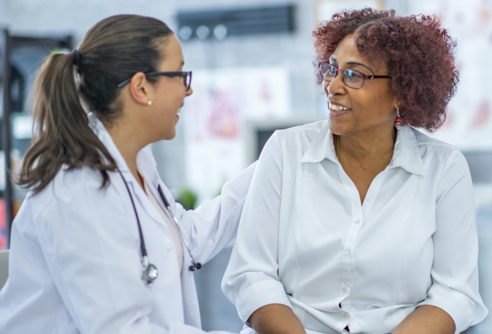 An African American woman smiles lightly as she discusses with the doctor sitting beside her. They are in a medical office.