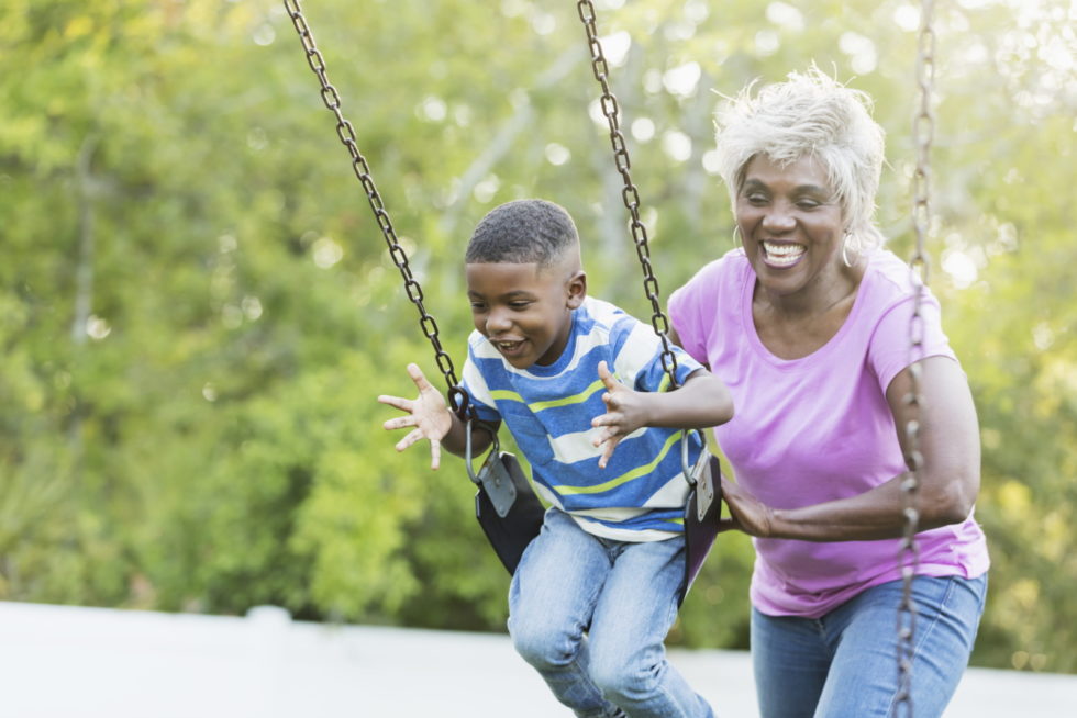 An elderly woman with peripheral artery disease watches her grandson happily swinging on a playground swing.