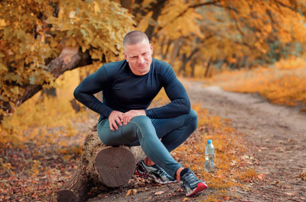 A young athletic jogger in black tight fitting sportswears and sneakers sits on a log, feels a strong pain in the muscle after cramping on a autumnall forest background.